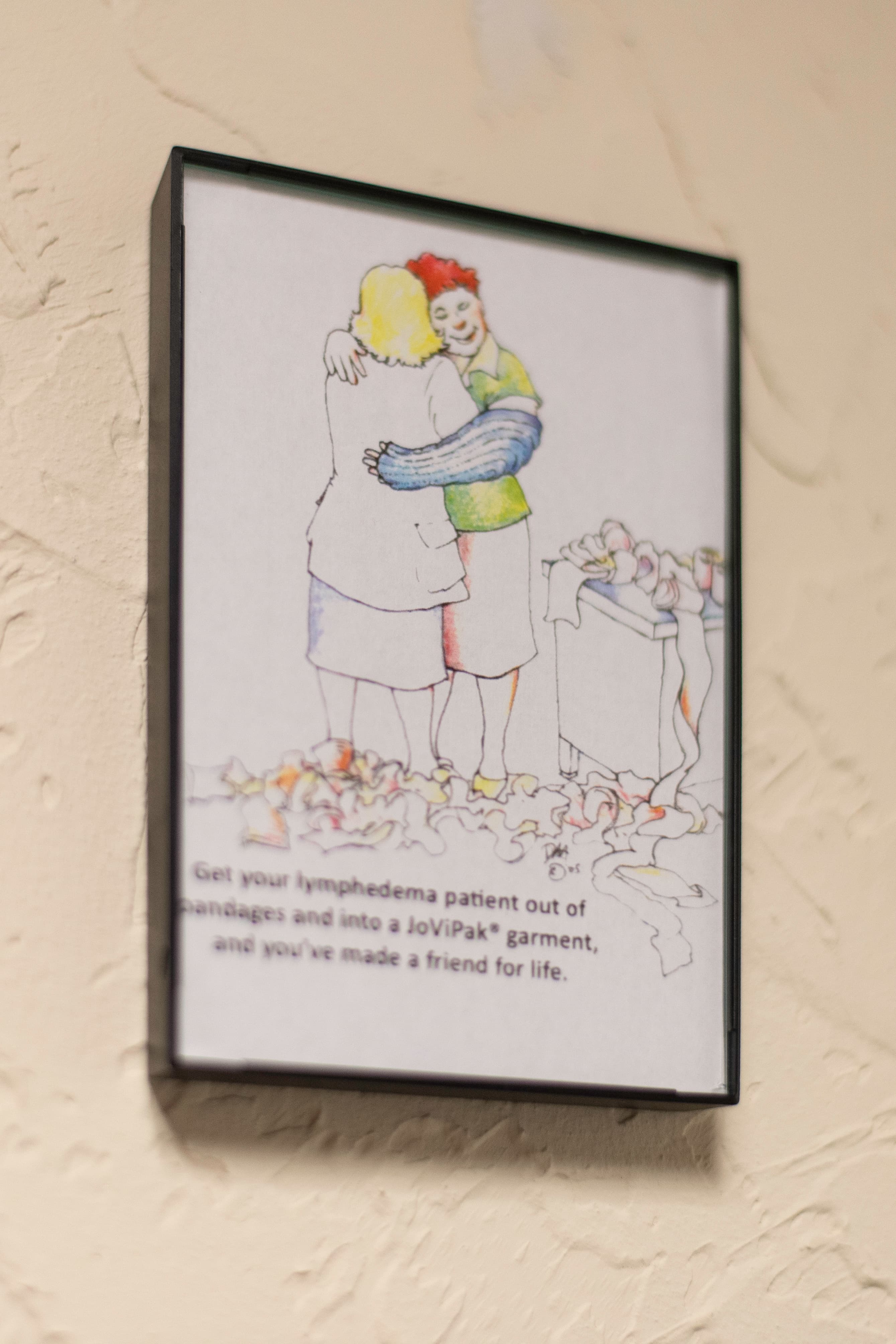 Photo of a framed comic depicting a clinician and lymphedema patient embracing each other next to an overflowing pile of bandages with the caption "Get your lymphedema patient out of bandages and into a JoViPak® garment, and you've made a friend for life."