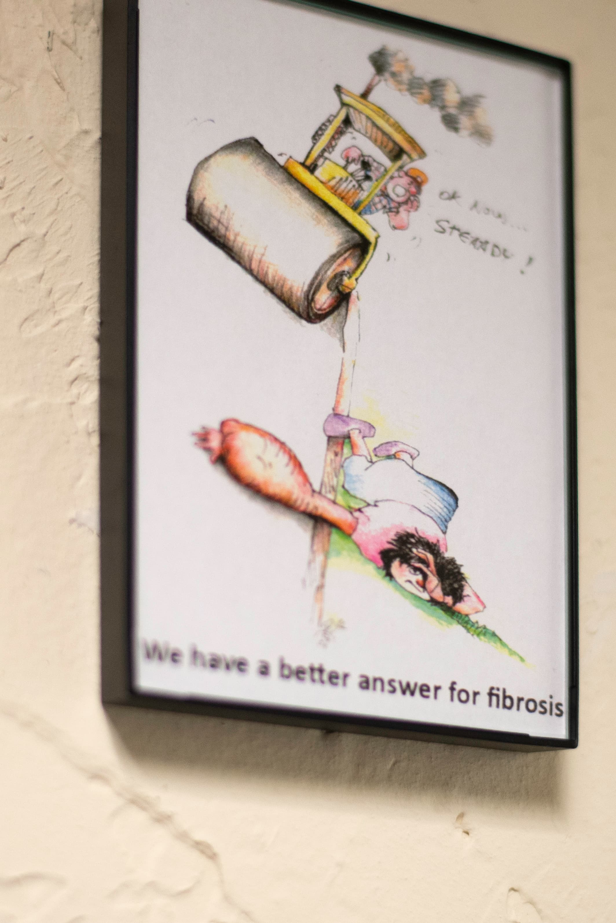Photo of a framed comic depicting a person lying prone next to a stretch of road with an edematous arm held on the road with a steam roller approaching, the driver of which is shouting "Ok now... steady!" The cartoon is captioned "We have a better answer for fibrosis."